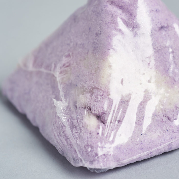 a close up of a purple pyramid shaped bath bomb wrapped in plastic packaging